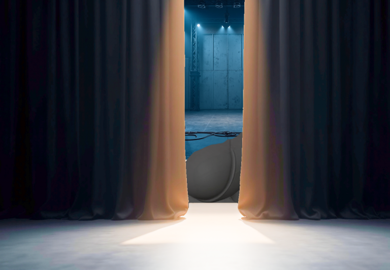 A dimly lit view from an audience, looking toward a stage where a thick black curtain is slightly open, revealing part of a large mechanical ride in spotlight and haze.